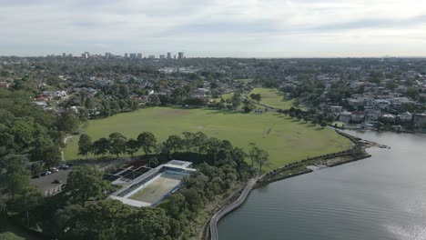 aerial fly over small town by a lush green forest and a with massive empty grass sport fields next to a lake in the morning with cloudy blue skies and empty swimming pool