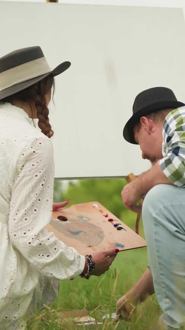 un hombre y una mujer se inclinan en un campo de hierba, preparándose para una sesión de pintura al aire libre. la mujer de blanco sostiene una paleta, mientras que el hombre de camisa a cuadros y sombrero recoge un tubo de pintura