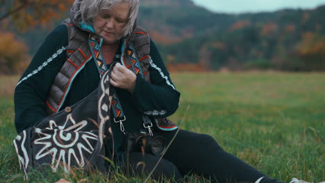 fat older woman with grey hair and camera looking for another lens in her bag while sitting on the grass in nature during a windy autumn day surrounded by colourful trees in slow motion