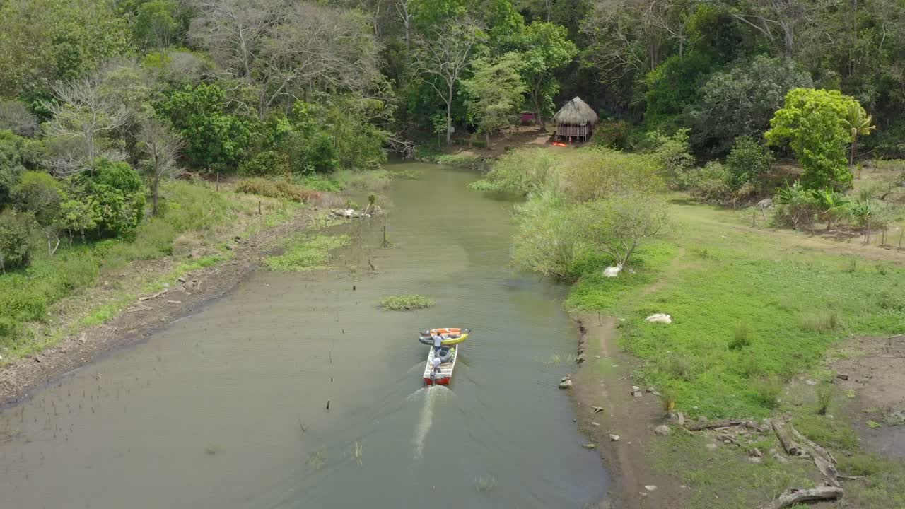 Aerial view of two men in a boat carrying two inflatable kayaks up a lake stream toward a bamboo hut, drone shot