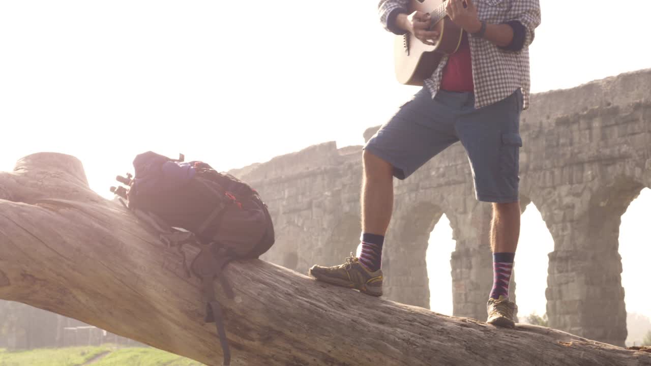joven aventurero viajero de pie en la parte superior de un tronco de tronco tocando la guitarra cantando frente a las antiguas ruinas del acueducto romano en el parque degli aquedotti en roma al amanecer en cámara lenta