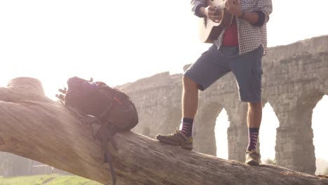 young man adventurer traveler standing on top of a log trunk playing guitar singing in front of ancient roman aqueduct ruins in parco degli acquedotti park in rome at sunrise slow motion