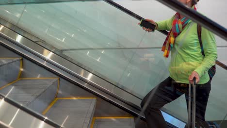 primer plano lateral de una mujer subiendo una escalera mecánica con un teléfono inteligente en la mano y equipaje rodante