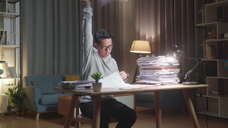 ein asiatischer mann, der papier wirft und kopfschmerzen hat, während er zu hause hart mit dokumenten arbeitet