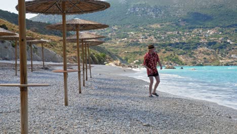 Hombre-Vestido-Con-Sombrero-Y-Traje-Floral-De-Playa-De-Verano-Tirando-Piedras-Al-Mar-En-La-Playa-De-Agia-Kiriaki,-Zola,-Cefalonia,-Grecia