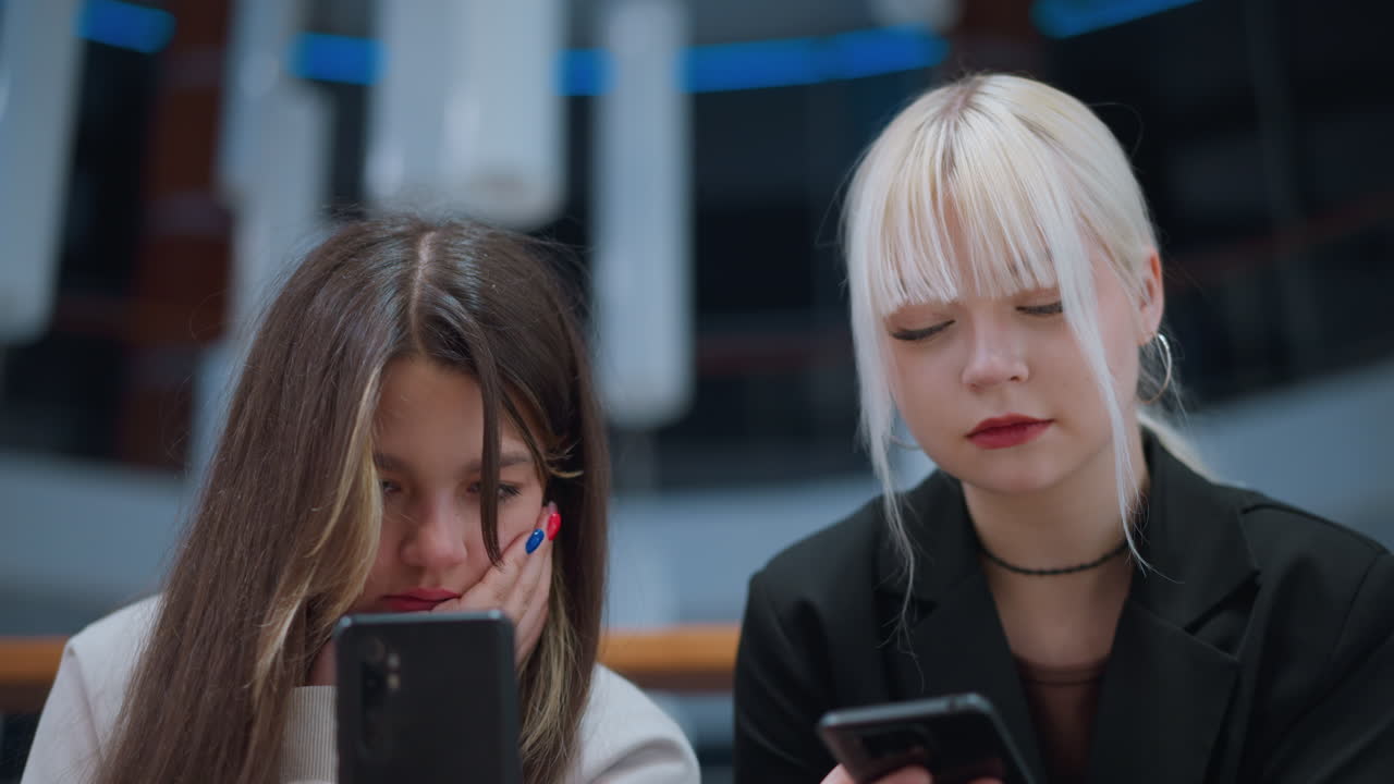 Close up of sisters focusing on phone while chatting indoors with serious expressions reflecting youth lifestyle, social media culture, digital communication and technology use in modern environment