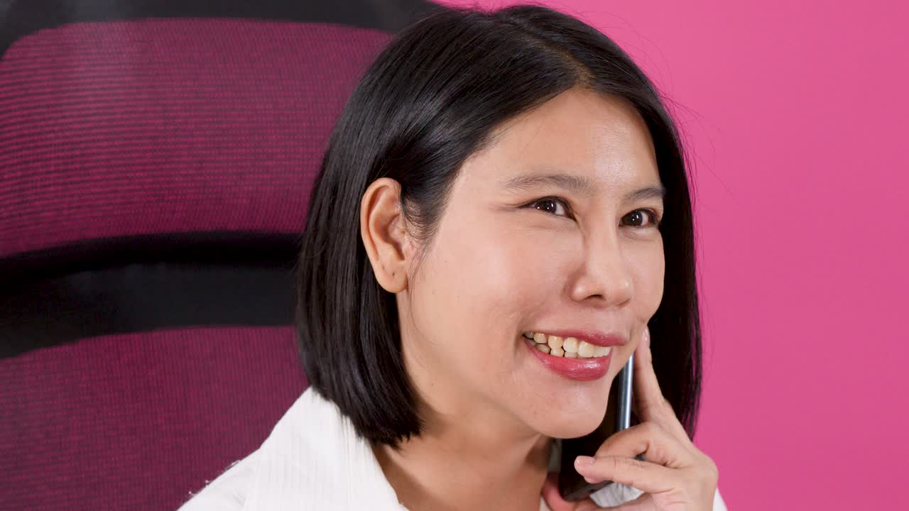 ピンクの背景に寄りかかりながら、スマートフォンで話す陽気なアジア人女性。