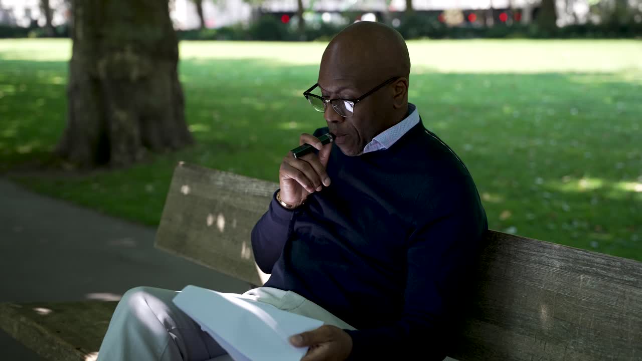 A man sits on a park bench in the shade, reading a white paper and smoking an e-cigarette, illustrating relaxation and leisure