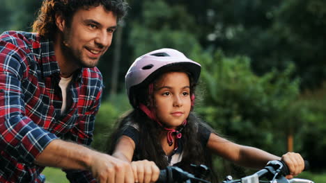 Closeup.-Portrait-of-a-little-girl.-Her-dad-is-teaching-her-to-ride-a-bike.-He-supports-her.-Moving-camera.-Blurred-background