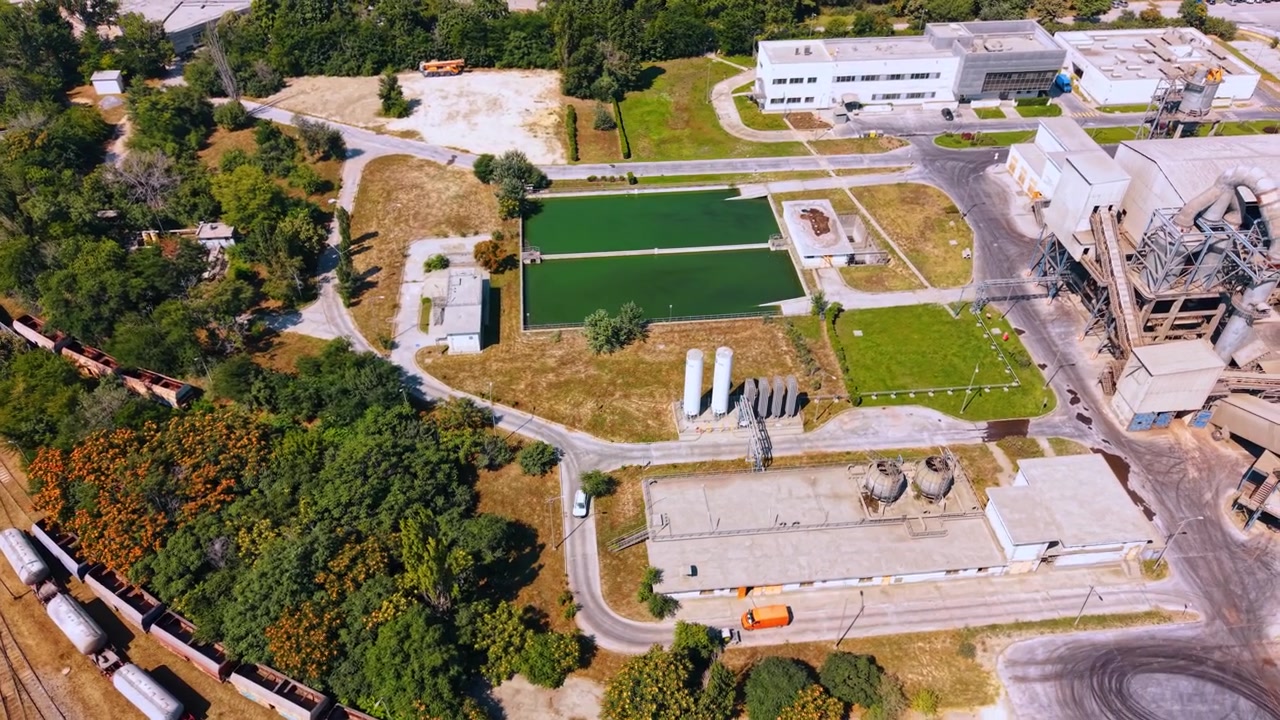 Bacini idrici industriali accanto a un piazzale ferroviario merci. Vista dall'alto di due stagni rettangolari verdi, edifici di servizio, strade, serbatoi, alberi e vagoni merci fermi.