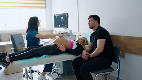 pregnant woman lies on the bed and doctor moves ultrasound device by her big belly. excited husband sits beside looking at the monitor.