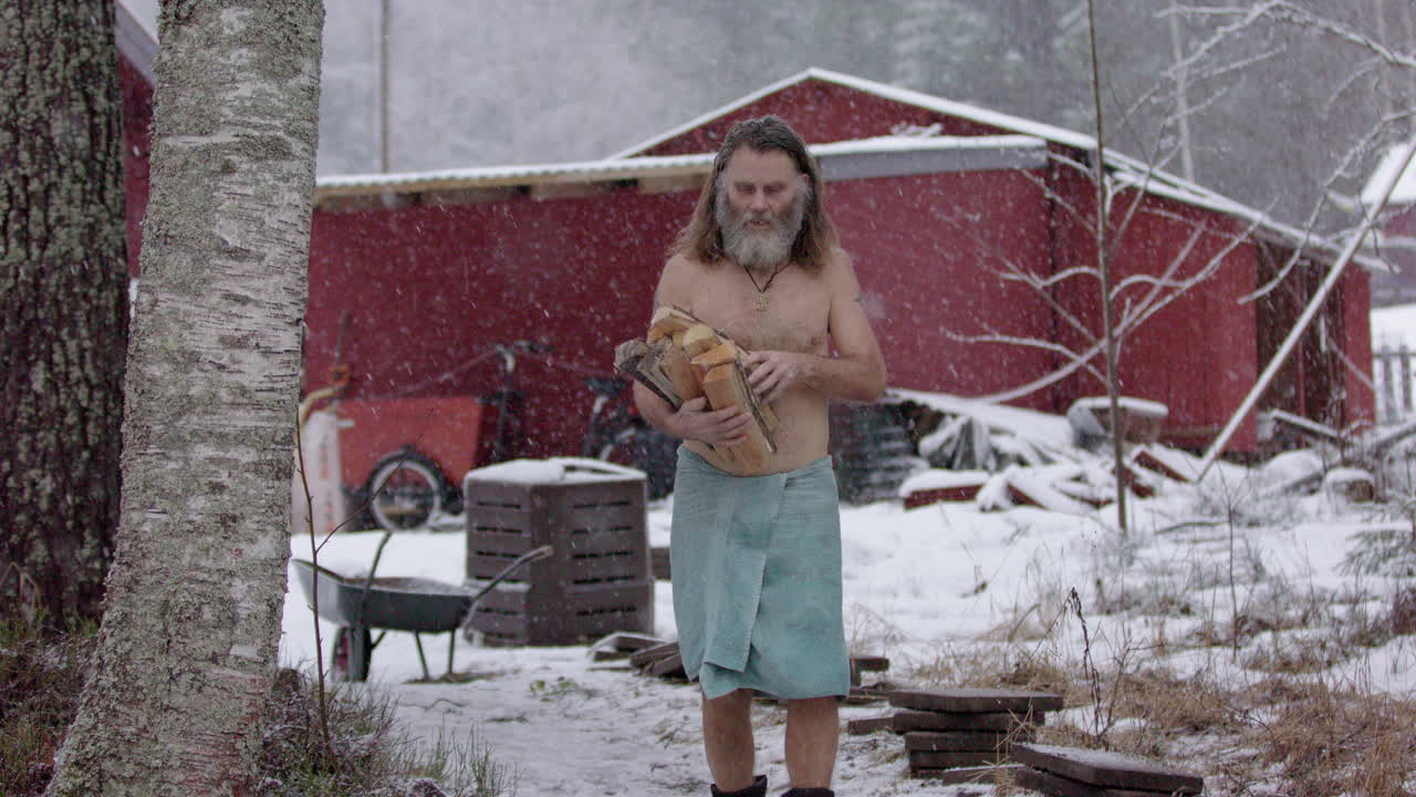 Bare chested viking man carries bundle of chopped firewood to sauna in snowfall