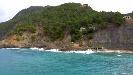 Wellen,-Die-An-Die-Klippen-Schlagen,-Und-Ein-Strand-Mit-Einem-Grünen-Hügel-Im-Hintergrund