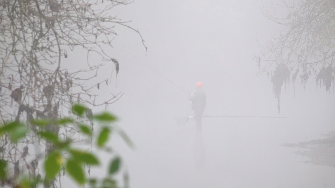 방수 바지와 밝은 주황색 모자를 쓴 어부가 얕은 강 옆에 있는 상륙 그물 옆에 서서 안개가 자욱한 이른 겨울 아침에 막대를 던집니다.