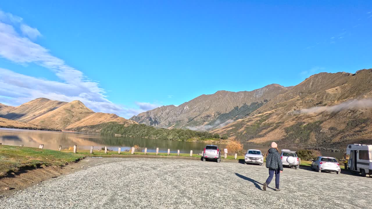 Individual walks across gravel car park toward vehicles, mountains, and lake under bright daylight