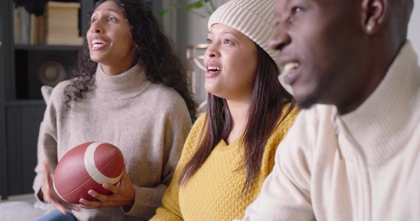 African American friends watching game on couch in living room, reacting to big play with football