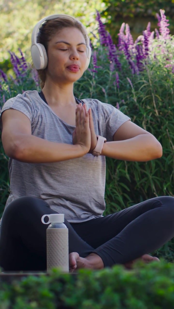 Vídeo vertical: Mulher com fone de ouvido respirando e movendo as mãos no jardim, áudio começando, com garrafa