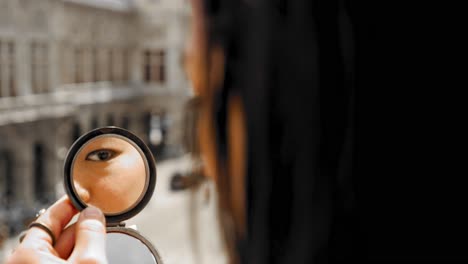 Beautiful-woman-putting-on-makeup-with-small-pocket-mirror-held-in-one-hand,-Vienna-Operhause-in-background