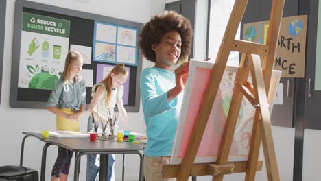 video of happy african american schoolboy at easel painting in diverse art class, copy space