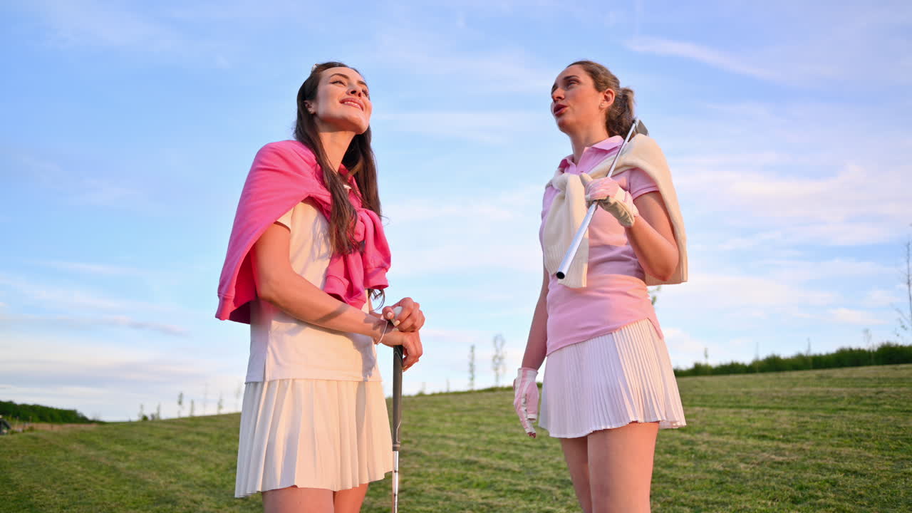 Two women dressed in white and pink clothes, holding golf clubs and talking on the course