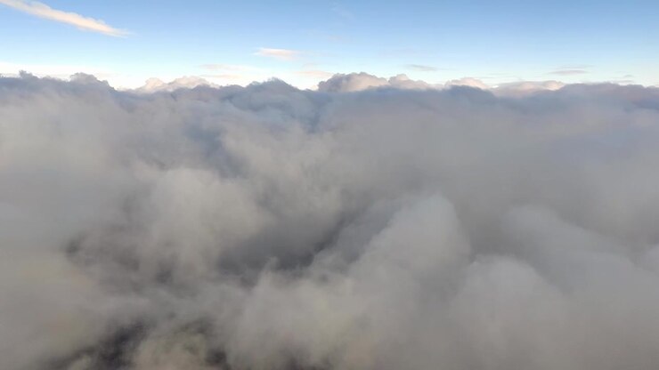Thick dense clouds fill the lower frame, with blue sky and bright sun above. Viewed from airplane window during flight