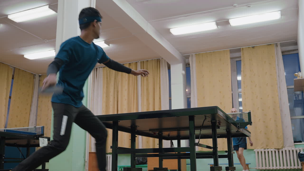 Young tennis player in blue shirt trains indoors with focus and determination while older coach observes, highlighting discipline, preparation, and training environment