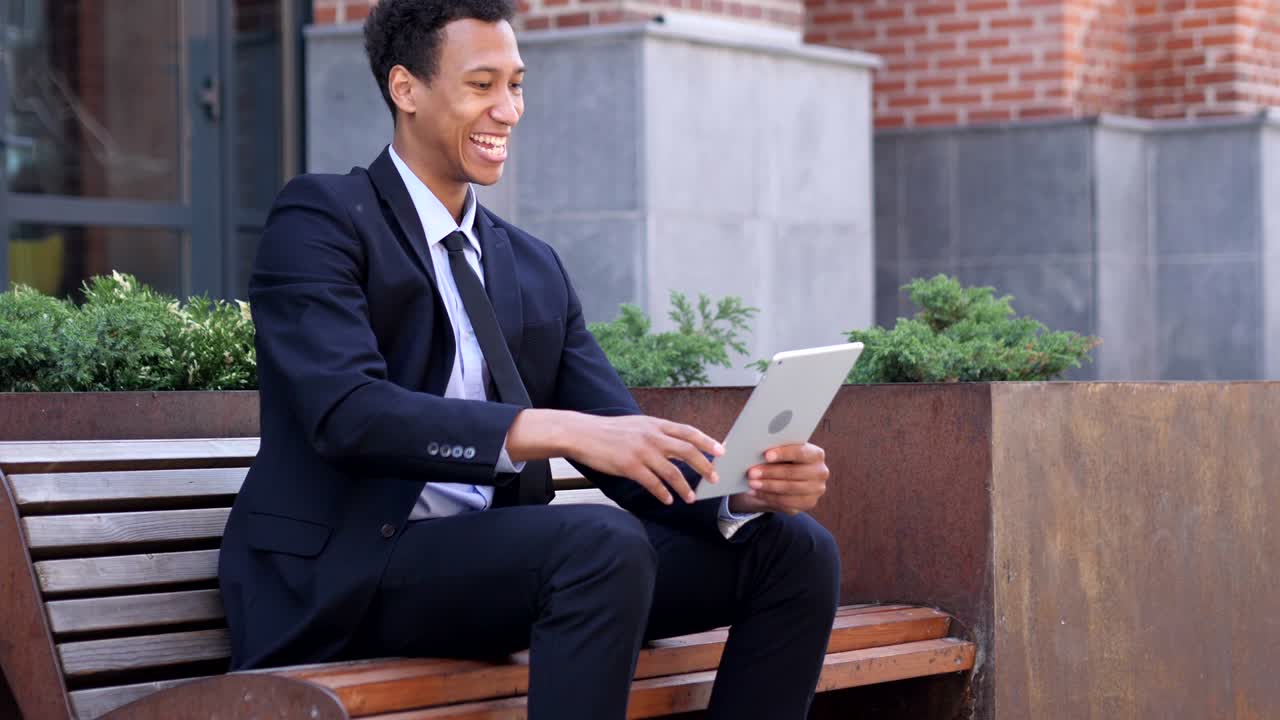 흥분 된 아프리카 사업가 가 벤치 에 앉아 있는 태블릿 에서 성공 을 축하 하고 있다