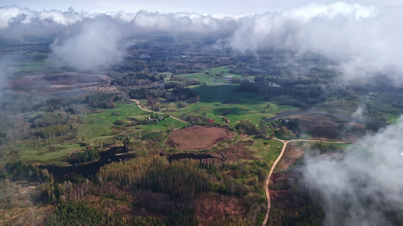Aerial above clouds of rural landscape with forests, green fields, and winding dirt roads