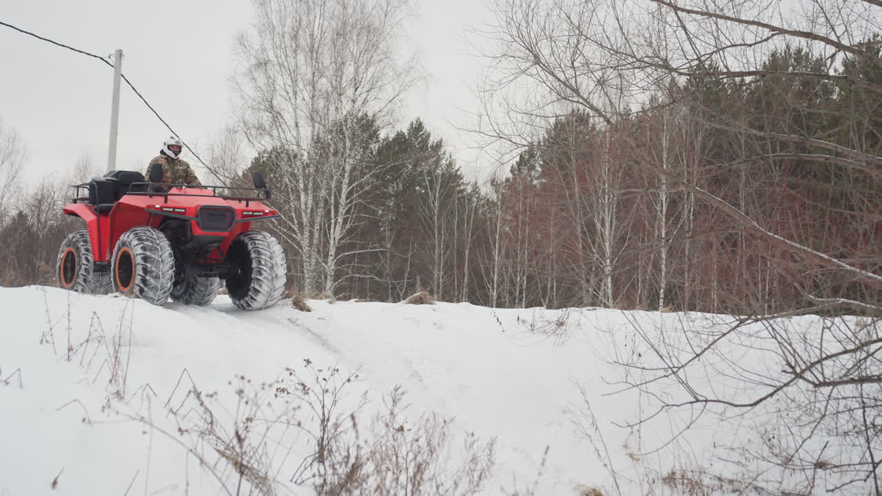Pilota caucasico su ATV rosso con giacca mimetica da neve e casco bianco che si lancia attraverso una foresta innevata. Grandi pneumatici chiodati sollevano neve polverosa. Salto dinamico e derapata su terreno ghiacciato, sotto un cielo coperto e tra betulle spoglie.