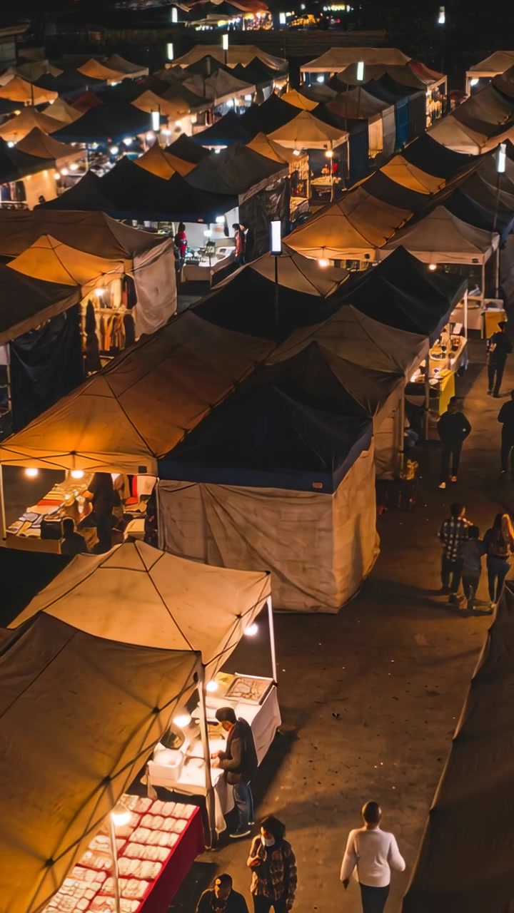 Vertical video: Panning drone showing lit canopy night market stalls, vendors serving buyers