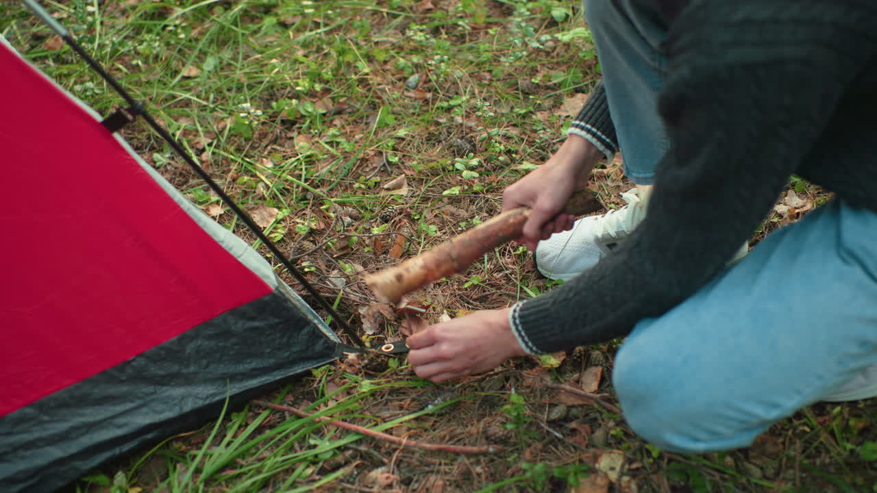 top down view of boy squatting near red tent securing tent corner by hammering metal peg into forest ground using thick wooden stick while wearing jeans sweater and sneakers