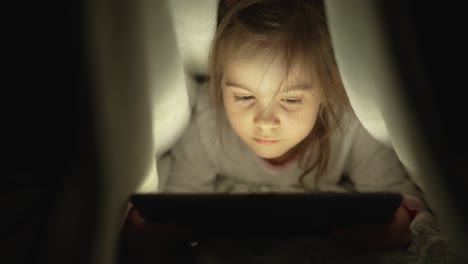 niño leyendo en la tableta bajo la manta por la noche