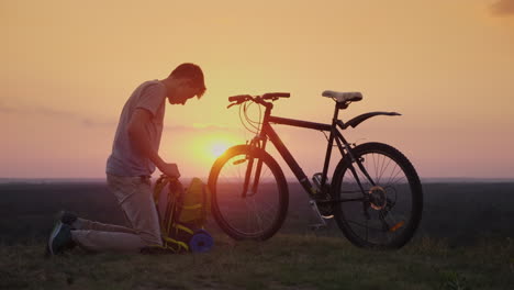 un joven pone cosas en una mochila se sienta junto a la bicicleta al atardecer 4k video de 10 bits