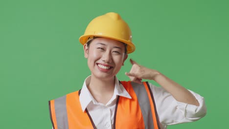 close up of asian female engineer with safety helmet smiling to camera and making call me gesture while standing in the green screen background studio