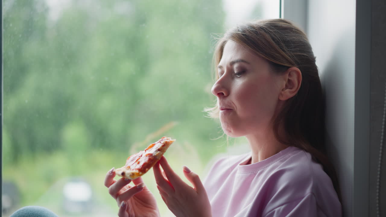 Woman seated by window enjoying slice of pizza with peaceful expression, wearing light purple shirt, soft daylight illuminating her face through glass with blurred green outdoor background