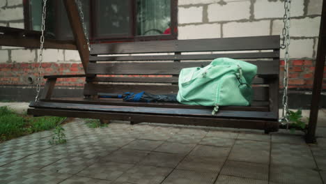 mint-colored bag and blue umbrella resting on wooden swing bench outside, with background showing partially constructed building, greenery, and interlocked pavement