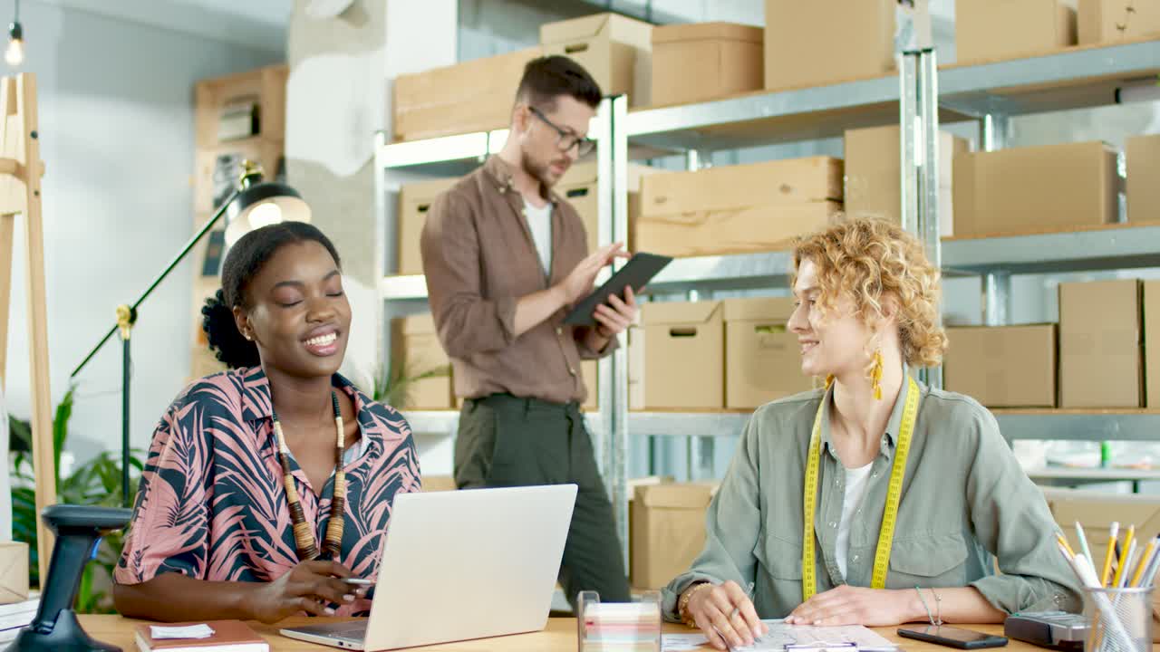 equipo de diseñadores multiétnicos escribiendo en una tableta, navegando en una laptop y hablando por teléfono y hablando entre ellos en una tienda de ropa de moda