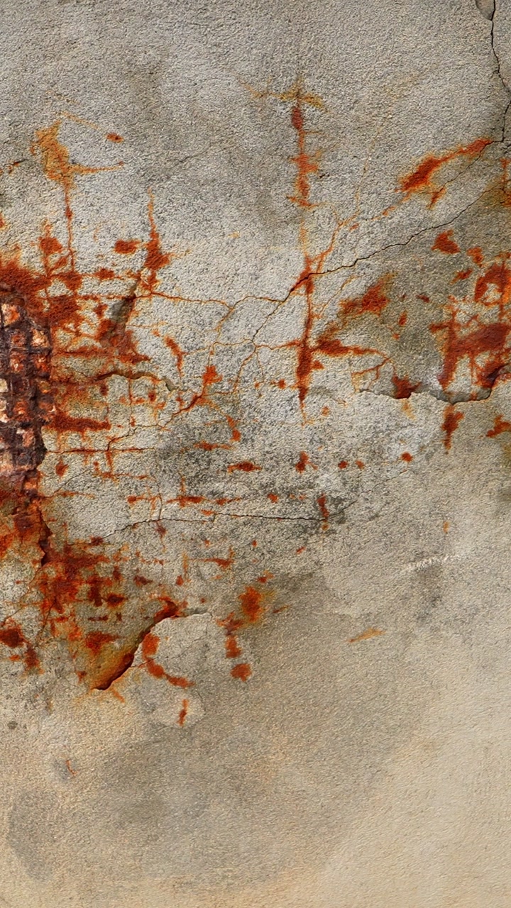 Vertical close-up of corroded wall render with exposed metal mesh and orange staining, showing water intrusion, structural decay, and deteriorating building surface.