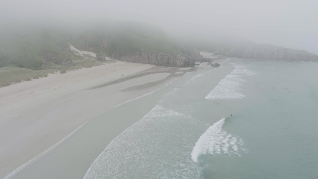 Slow drone reveal shot of surfers, big waves and misty weather on Ceannabeinne Beach on the Scotland North Coast 500 Route which is the North Sea