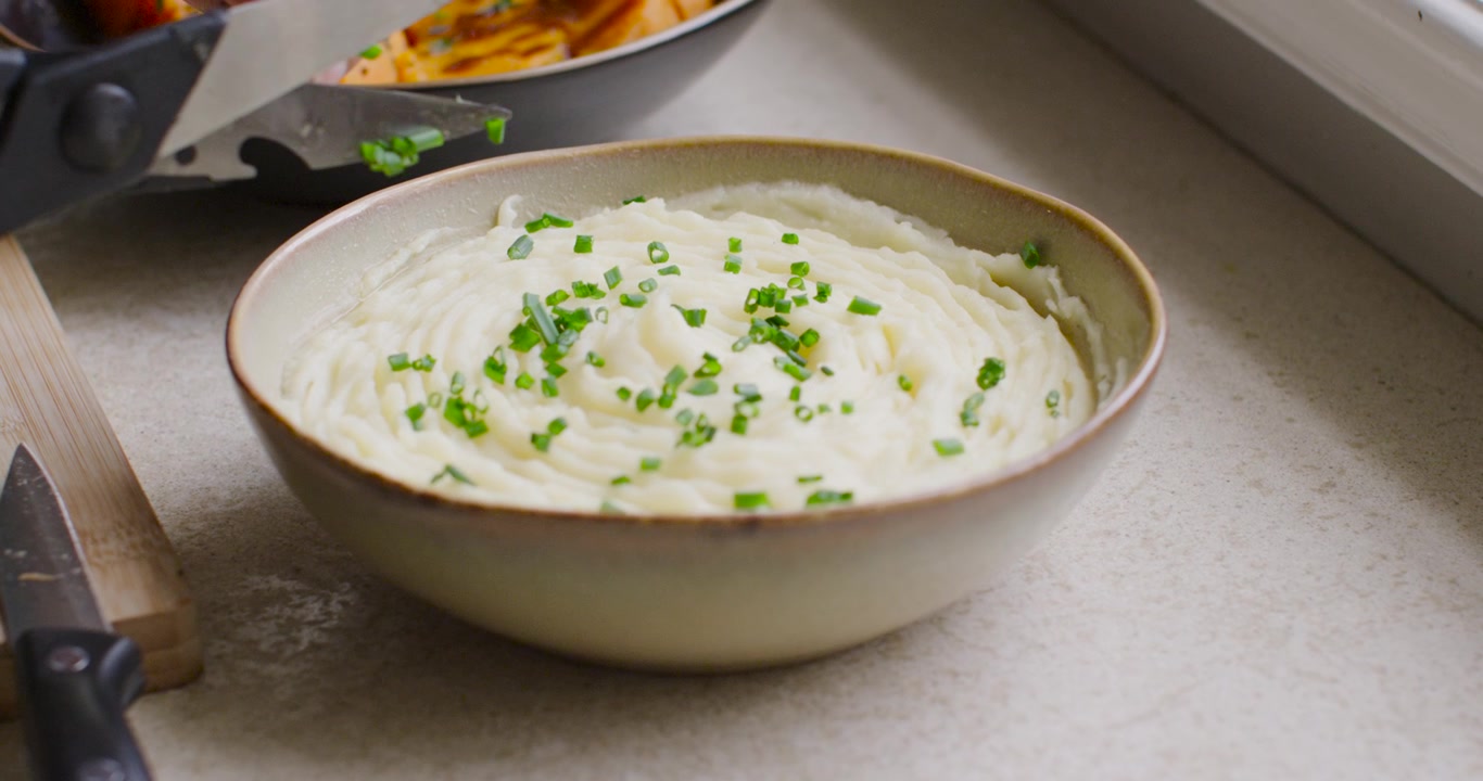 Mãos de cozinheiro com luva verde entrando pela esquerda, usando tesoura para cortar cebolinha e emplatar purê de batatas e tubérculos
