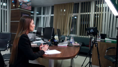 long-haired brunette lady sits at the desk in the office. woman focused on camera speaks gesturing with her hands. side view. blogging concept.