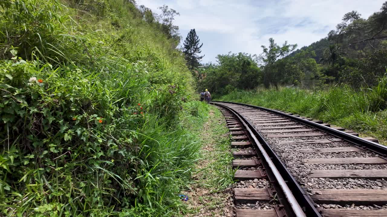 POV Walking beside railway track curves through the verdant landscape of Ella, Sri Lanka. The lush green surroundings and clear sky create a serene and peaceful atmosphere.