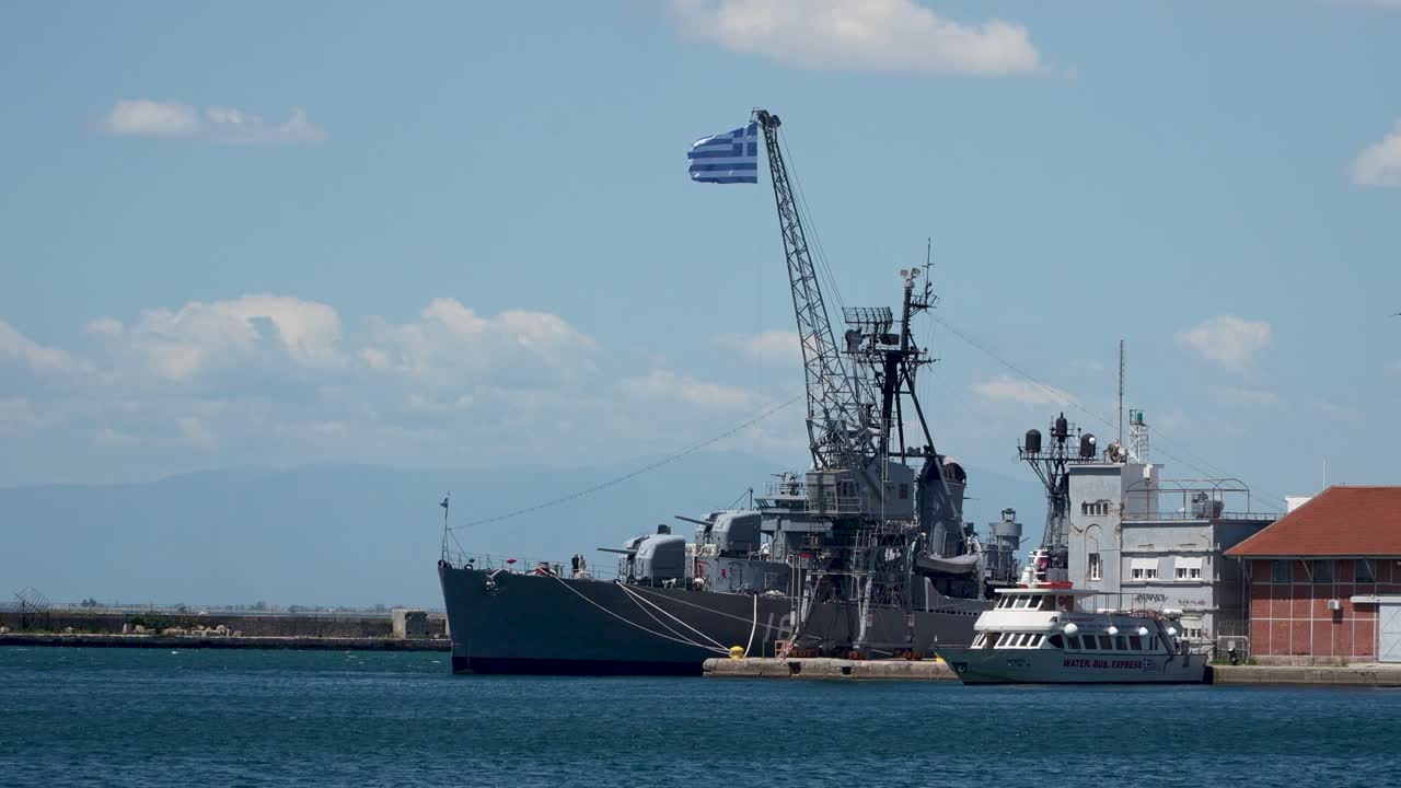A Greek naval warship is docked at a harbor beside a "Water Bus Express" vessel. A large Greek flag flies atop a crane near the pier, with nearby port facilities and calm blue waters