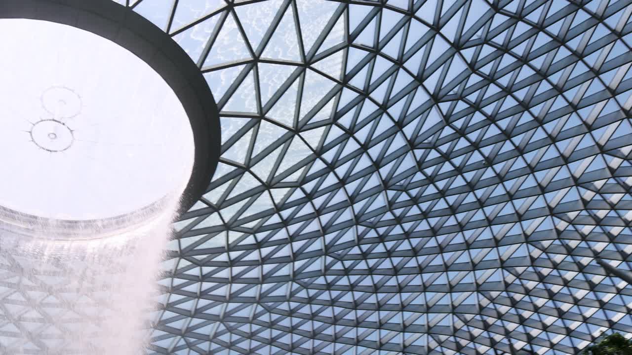 Camera tilts upward from tropical trees to reveal a dramatic indoor waterfall cascading from a geometric glass dome, bathed in natural daylight