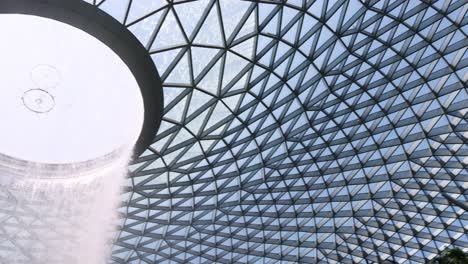 camera tilts upward from tropical trees to reveal a dramatic indoor waterfall cascading from a geometric glass dome, bathed in natural daylight