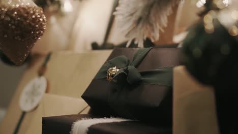 static shot of a christmas present wrapped with black wrapping paper and green ribbon and a bell underneath a christmas tree with other presents