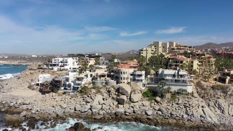 a drone glides laterally over the opulent coastal homes of cabo san lucas, capturing the essence of luxury against the backdrop of crashing waves