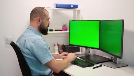hombre barbudo bebiendo agua de un vaso sentado en una mesa frente a dos pantallas de teclas de croma verde en primer plano 4k.
