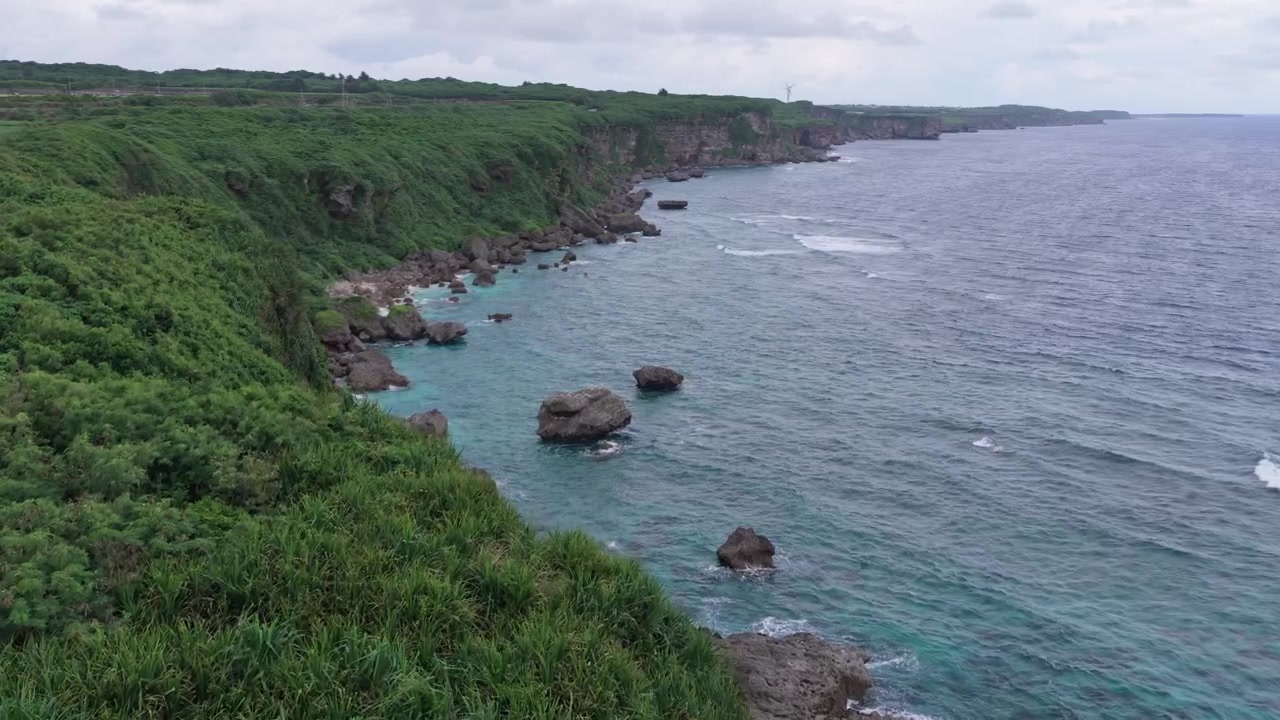Rekaman udara horizontal bergerak maju di atas garis pantai batu kapur terjal Tebing Muiga dengan Kincir Angin Energi yang jauh, Miyakojima, Okinawa, Jepang.