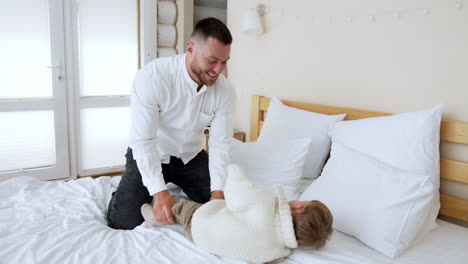 caucasian father throws his little son on the pillows playfully. loving dad kisses a baby. woman stands behind leaning on the wall.